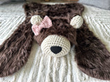 Slightly elevated front view of the brown faux fur bear rug flat on a cream carpet. Features a pink crochet bow attached below the right ear, on the left side of the photo. Cozy accent rug for a woodland theme nursery.