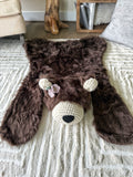 An angled front view looking down at the plush faux fur bear rug. Highlights the handmade light purple and green crochet flower accessory stitched to the bear's head on a cream carpet. Cute woodland theme nursery mat.