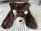 Slightly elevated front view of the brown faux fur bear rug on a cream carpet. Purple and green crochet flowers are attached to the right ear, shown on the left side of the photo. Perfect woodland nursery floor decor.
