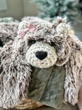 A plush bear lovey with brown and beige tipped faux fur, black safety eyes, and a pink crochet bow, draped over a green blanket.