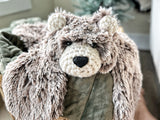 A plush bear lovey with brown and beige tipped faux fur and black safety eyes, draped over a green blanket.