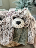 A plush bear lovey with brown and beige tipped faux fur, black safety eyes, and purple and green crochet flowers, draped over a green blanket.