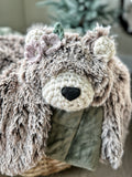 A plush bear lovey made with brown and beige tipped faux fur and purple and green crochet flowers by its ear.