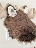 Close-up of a brown faux fur moose wall mount showing the plush fur texture, black safety eyes, and purple and green crochet flowers.