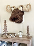 A front left side view of a brown faux fur moose wall mount hanging above a side table with a snow globe and two wooden tree sculptures.