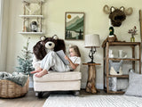A girl sitting on a white rocking chair with a faux fur grizzly bear rug draped over the back, in a woodland bedroom featuring a plush moose wall mount.