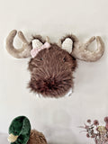 Front view of a brown faux fur moose wall mount with a pink crochet bow below its right ear, on a white plaque.