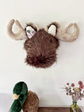 Front view of a brown faux fur moose wall mount with purple and green crochet flowers below its right ear.
