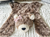 Overhead front angle view of the cappuccino faux fur bear rug. A pink crochet bow is attached to the right ear, appearing on the left side of the photo. Placed on a beige carpet as cozy woodland nursery decor.