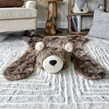 Plush cappuccino faux fur bear rug with cream crochet ears, a cream crochet muzzle, and a black nose, displayed flat on a beige carpet. Cozy woodland nursery decor by ClaraLoo.