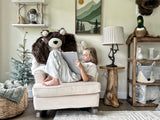 Close-up view of a young girl reading a book in a white rocking chair draped with the brown faux fur bear rug. Highlights the plush, soft texture of the brown bear mat in a cozy woodland nursery.