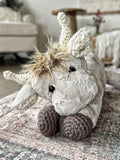 Plush highland cow in a kneeling position on a carpet. The highland cow toy is made with tan fur and has crochet horns, ears, and feet.