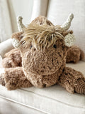 A large plush brown highland cow with cream crochet horns and brown crochet feet, sitting sprawled out on a white rocking chair.
