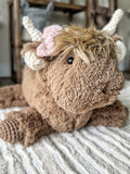 Front angled view of a plush brown highland cow with a pink crochet bow by its horn, sitting on a beige and white striped rug.