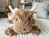 Close-up headshot of a plush brown highland cow with soft minky fur, cream crochet horns, and a pink crochet bow.
