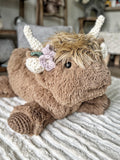 Front right side view of a plush brown highland cow with purple and green crochet flowers by its horn, sitting on the carpet.