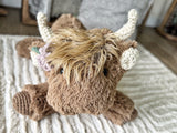 Close-up headshot of a plush brown highland cow with purple and green crochet flowers attached below its horn.
