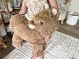 A child holding a large plush brown highland cow with soft minky fur and cream-colored crochet horns, sitting on a white and cream rug.