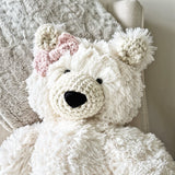 A close-up view of a white plush polar bear stuffed animal with black safety eyes and a light pink crochet bow attached underneath its right ear on the left side of the photo.