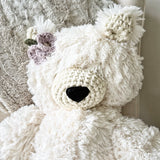 A close-up view of a white plush polar bear stuffed animal with cream colored crochet accents and purple and green crochet flowers attached underneath its right ear on the left side of the photo.