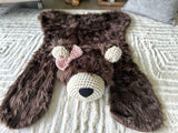 Slightly elevated head-on view of the plush brown faux fur bear rug on a cream carpet. Showcases the symmetrical cream crochet muzzle, ears, and black nose details. Woodland nursery theme floor mat.