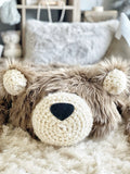Close-up view focusing on the head of the brown faux fur bear rug with cream crochet ears and a matching cream crochet muzzle. The plush bear mat is resting flat on a soft, fluffy cream-colored carpet.