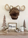 A faux fur brown moose wall mount with cream crochet ears and tan antlers on a white plaque, hanging above two wooden tree sculptures.