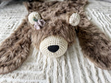 Front angle view of the cappuccino faux fur bear rug head. Light purple and green crochet flowers are attached below the right ear, shown on the left side of the photo. Perfect animal floor mat for a woodland theme nursery.