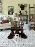 Wide view of a brown faux fur bear rug flat on a cream carpet. In the background, there is a white rocking chair with a brown pillow. Perfect design inspiration for a cozy rustic child's nursery.