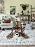 Cappuccino faux fur bear rug on a beige carpet in a woodland nursery. Background features a white rocking chair, wooden shelf with a crochet fox basket, plush moose wall mount, and a small table with a lamp.