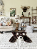 Wide view of a brown faux fur bear rug flat on a cream carpet. In the background, a young girl sits in a white rocking chair reading a book in a woodland themed bedroom. Perfect design inspiration for a cozy rustic child's nursery. Handmade nursery decor by ClaraLoo.