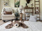 Wide lifestyle view of the cappuccino faux fur bear rug on a beige carpet in a woodland nursery. Background features a white rocking chair, a table with a black lamp, and a wooden ladder draped with an animal print blanket.